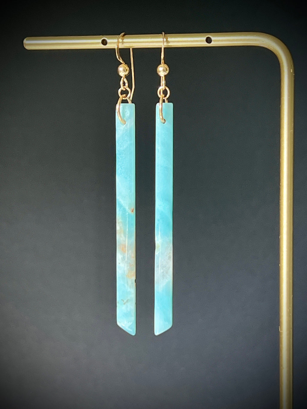 Amazonite long bar earrings hanging on a gold display frame against a dark grey background, highlighting the stone's bright teal and cream marbled pattern