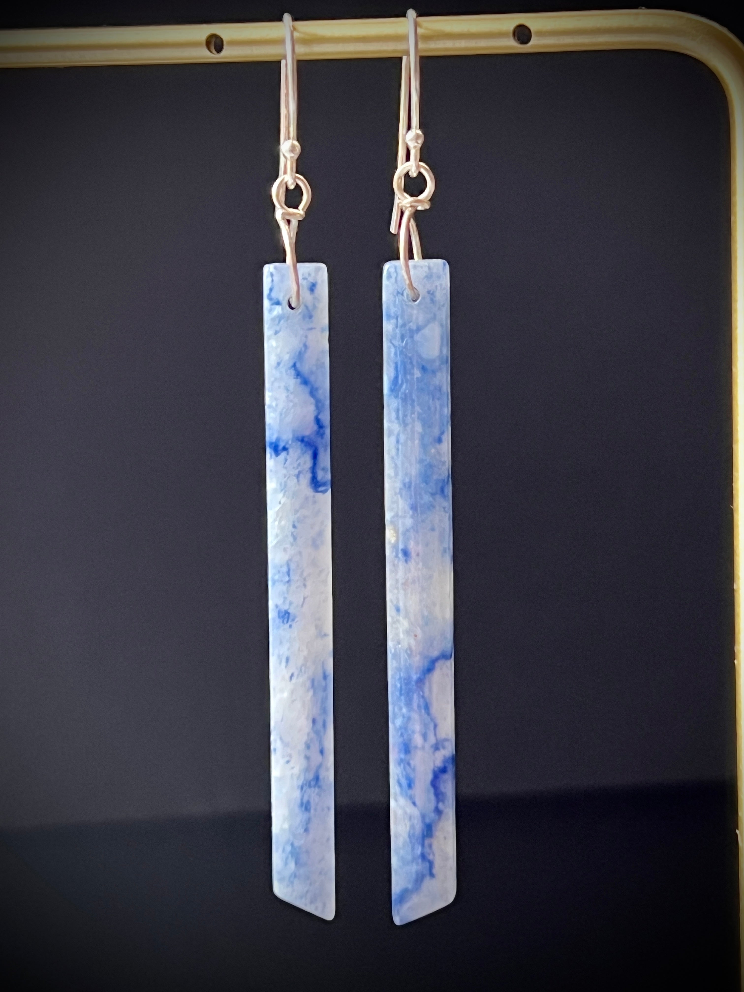 Blue aventurine long bar earrings hanging on a gold display frame against a dark gray background, highlighting the stone's blue and white marbled pattern