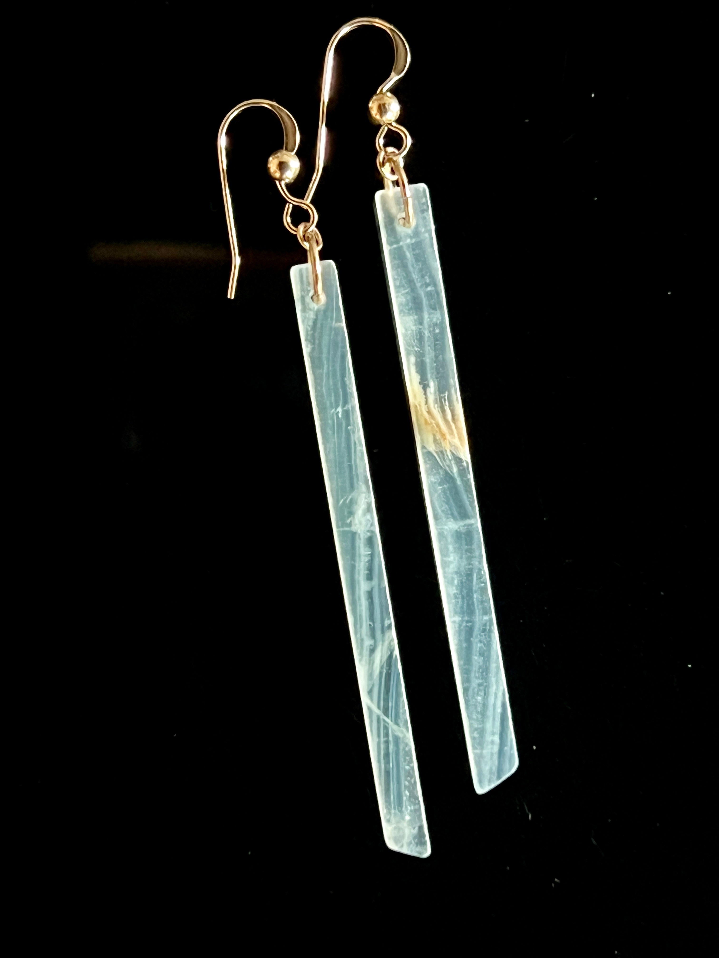 Pair of Lemurian calcite long bar drop earrings with 14K gold fill fish hook wires on a black background, showing pale blue-grey stone with warm gold inclusions