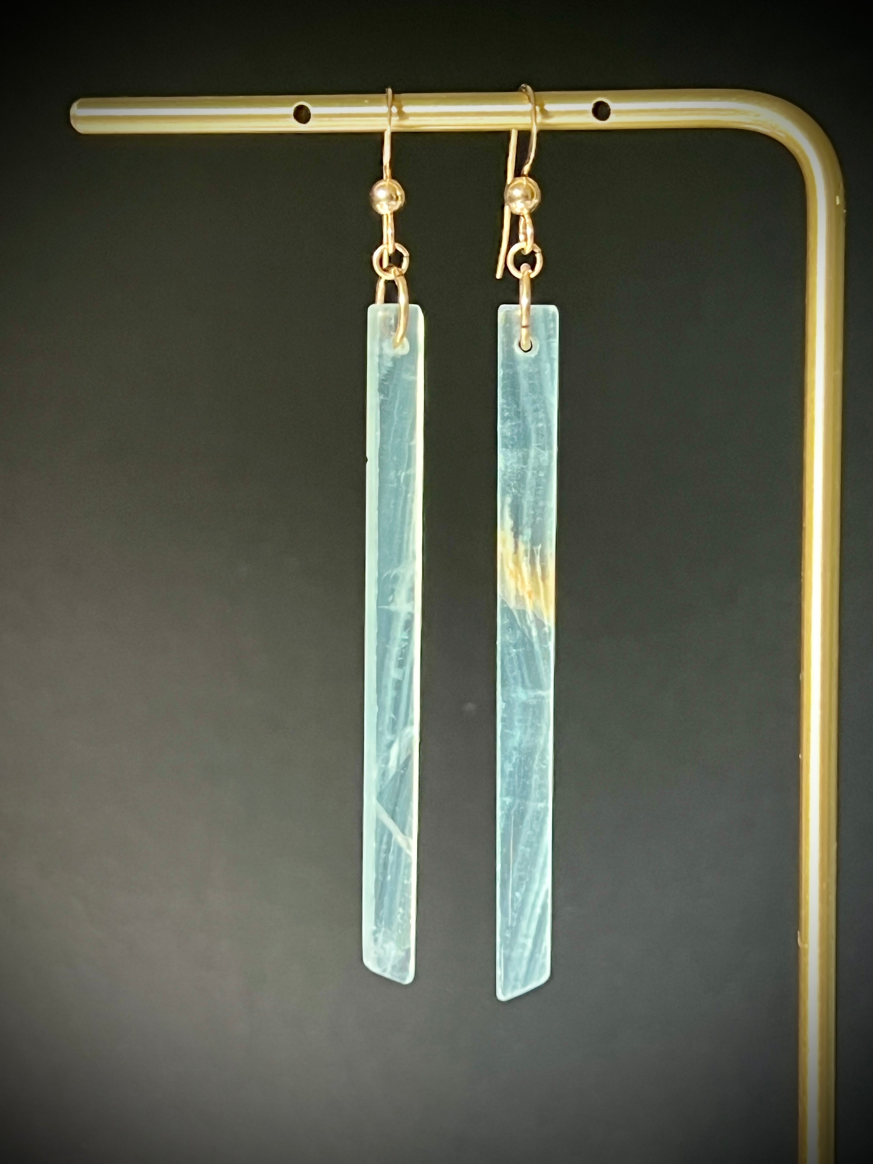 Lemurian calcite long bar earrings hanging on a gold display frame against a dark grey background, highlighting the stone's translucent pale blue tone and amber veining