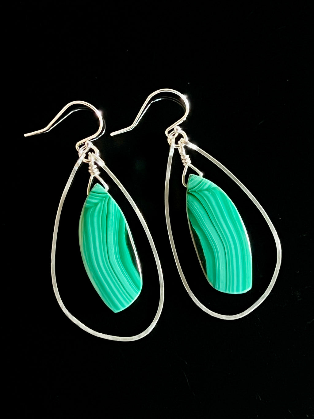 Pair of malachite and sterling silver freeform dangle earrings on a black background, showing vivid green banded marquise stones suspended inside open teardrop wire frames with fish hook wires