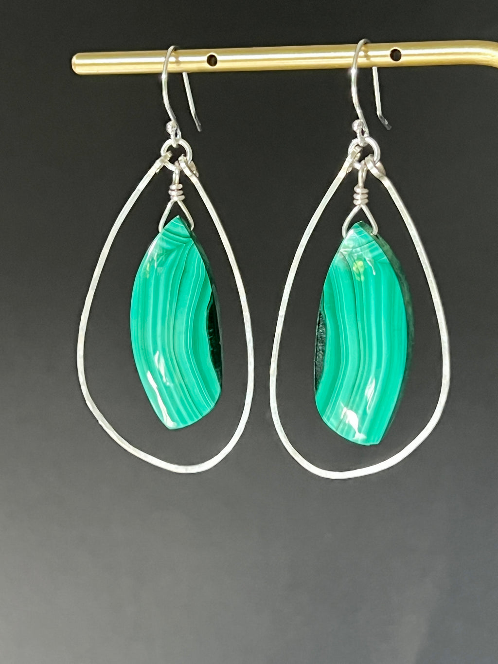 Malachite and sterling silver freeform dangle earrings hanging on a gold display bar against a dark grey background, highlighting the bright green and dark banding of the malachite stones