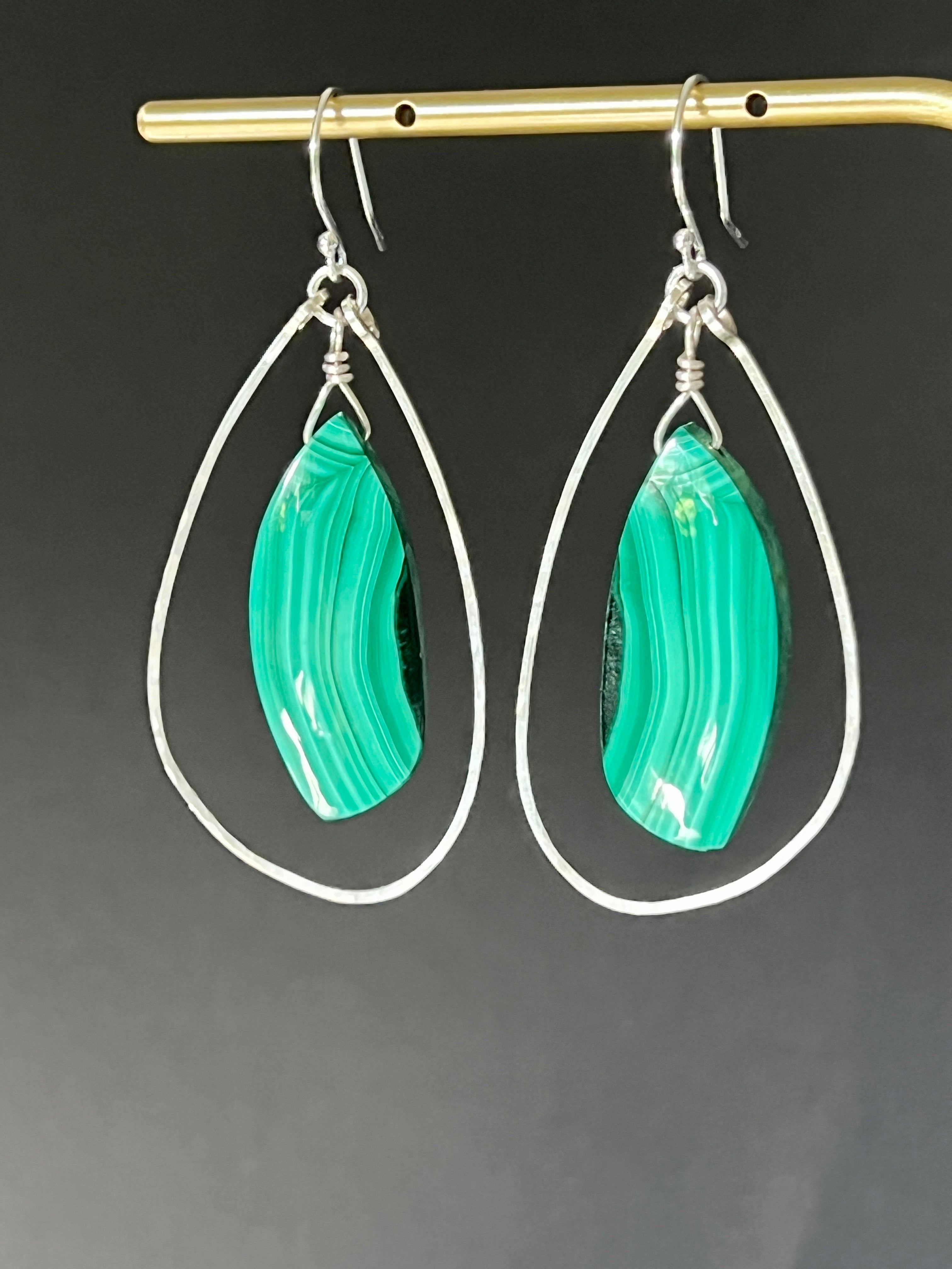 Malachite and sterling silver freeform dangle earrings hanging on a gold display bar against a dark grey background, highlighting the bright green and dark banding of the malachite stones
