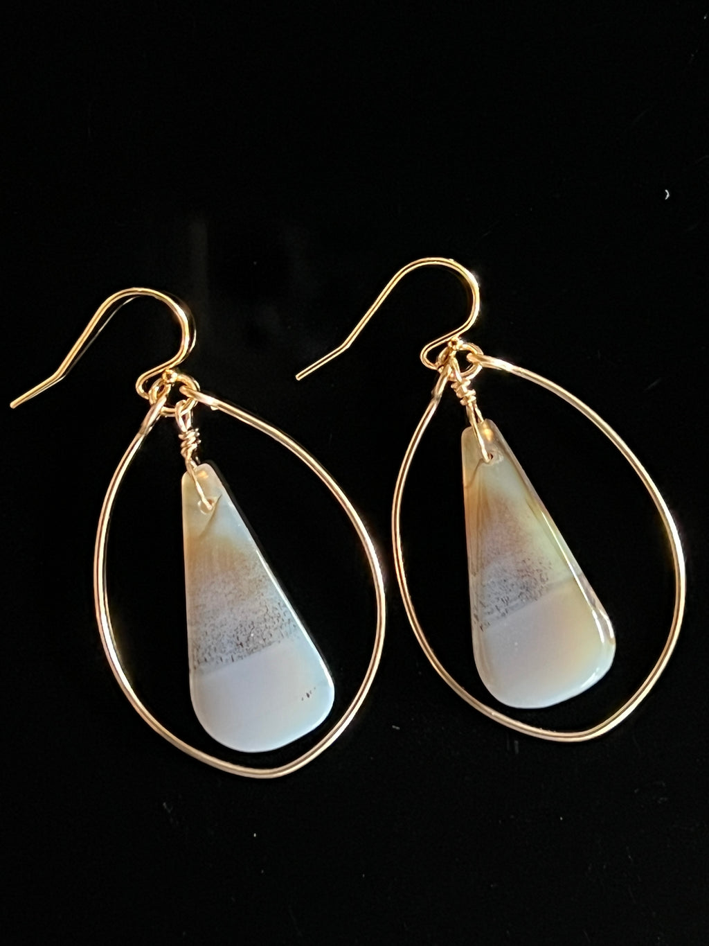 Pair of Montana agate dangle hoop earrings in 14K gold fill on black background, showing freeform white and warm sand stones with silvery banding inside oval gold wire frames by Studio MS Indie Jewelry