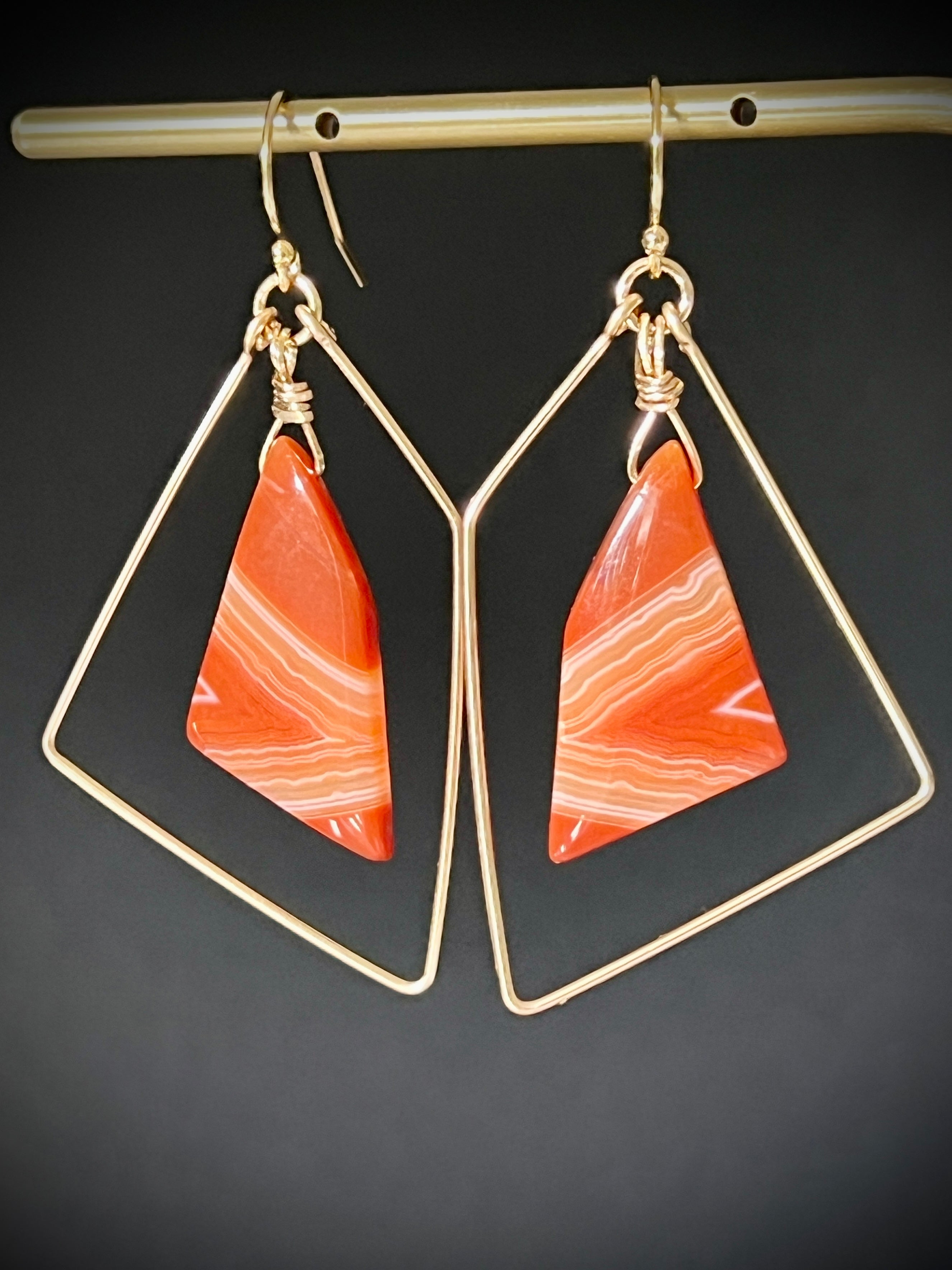 Pair of sardonyx freeform earrings in 14K gold fill hanging on a gold bar, dark gray background, showing bold orange banded stones in open geometric gold wire frames